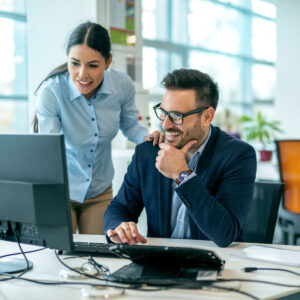 Standing female and seated male office workers smiling viewing a computer screen at a desk